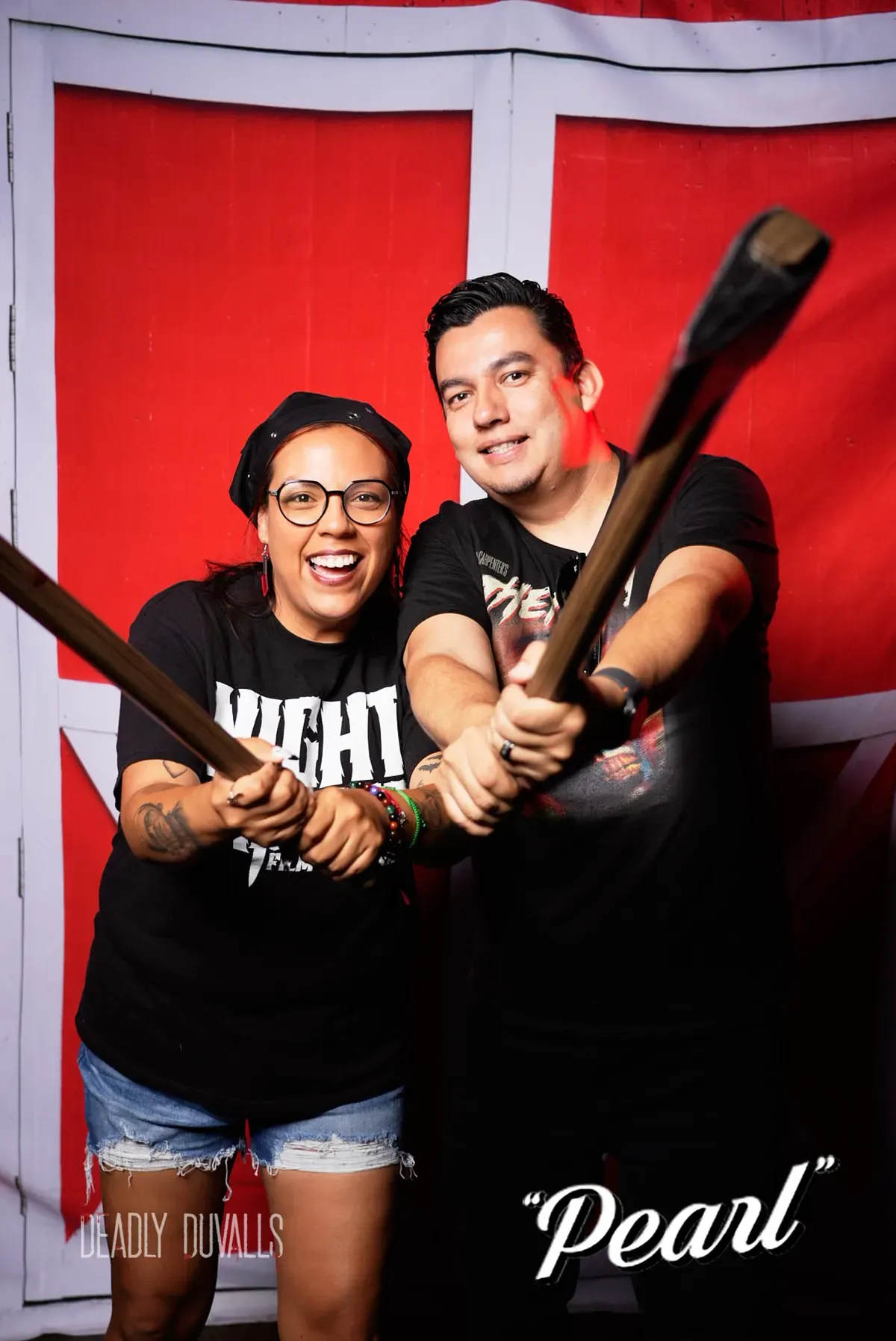 Two guests laughing with crossed prop weapons against the red barn-door backdrop at the Deadly Duvalls Pearl event photo booth