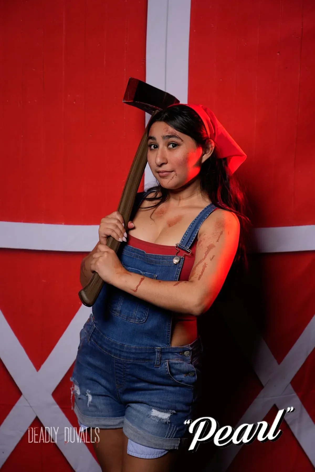 Guest posing as Pearl in denim overalls with a bloodied axe against a red barn-door backdrop at a Deadly Duvalls photo booth in Los Angeles