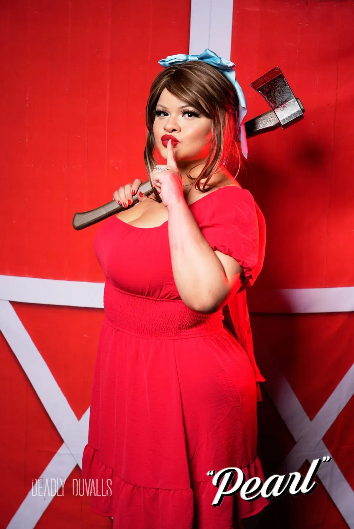 Guest in a red dress and blue hair bow holding an axe over her shoulder at the Pearl photo booth, red barn-door backdrop, Los Angeles