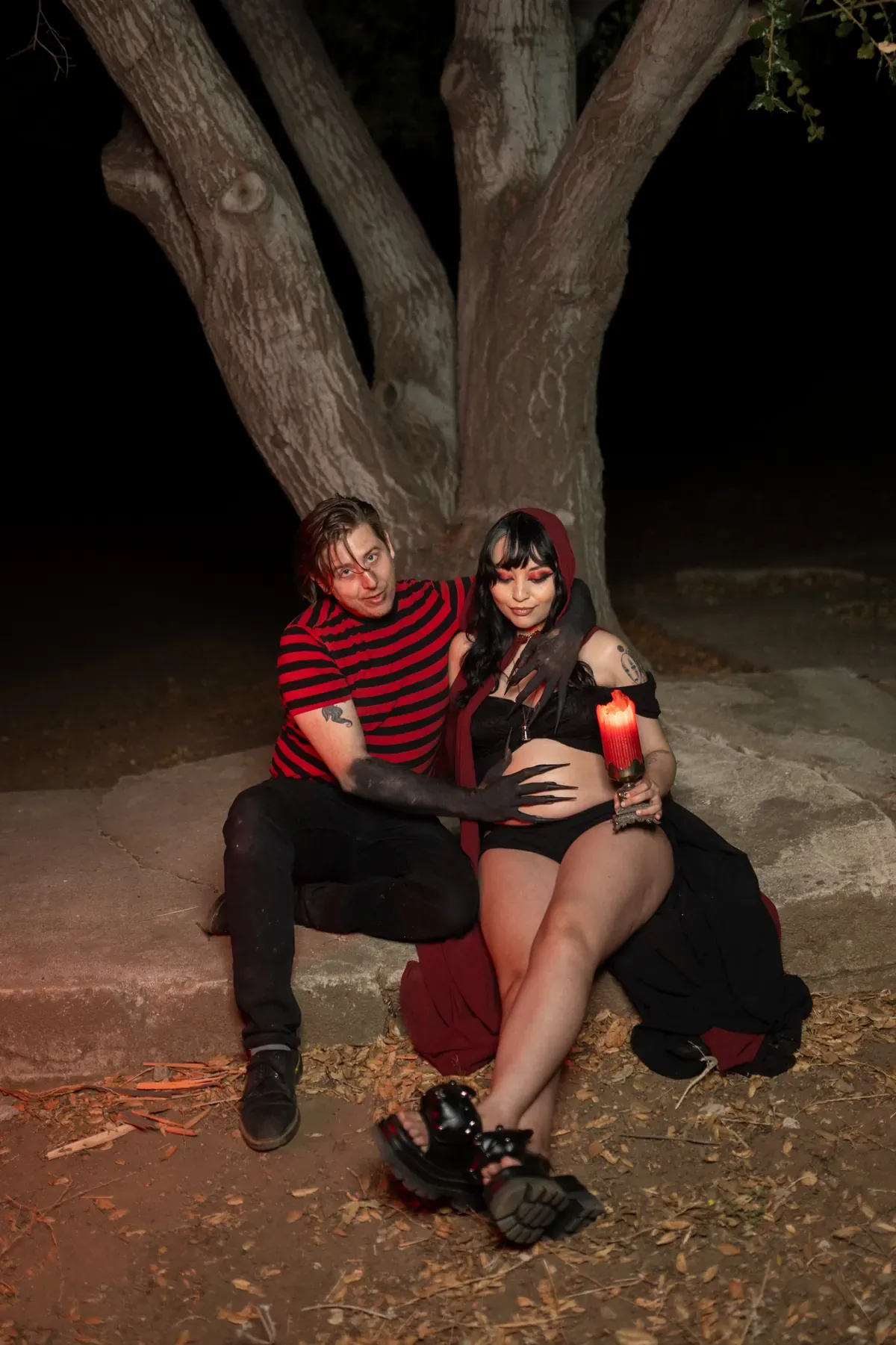 Expectant couple seated at the base of an oak tree at night, she in a red hood holding a lit red candle — gothic maternity couple portrait in Los Angeles