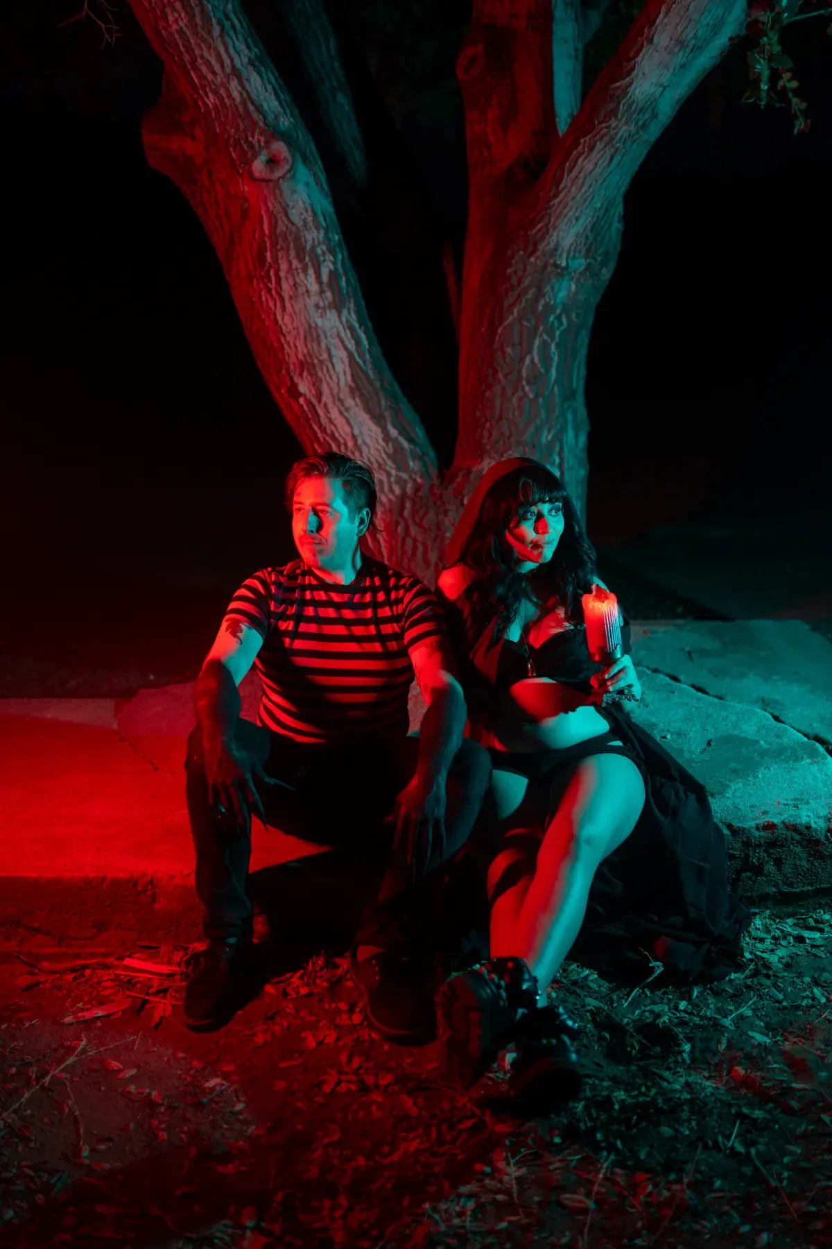 Expectant couple leaning into each other beneath an oak tree under red and teal gelled light — spooky maternity couple portrait by Deadly Duvalls in Los Angeles