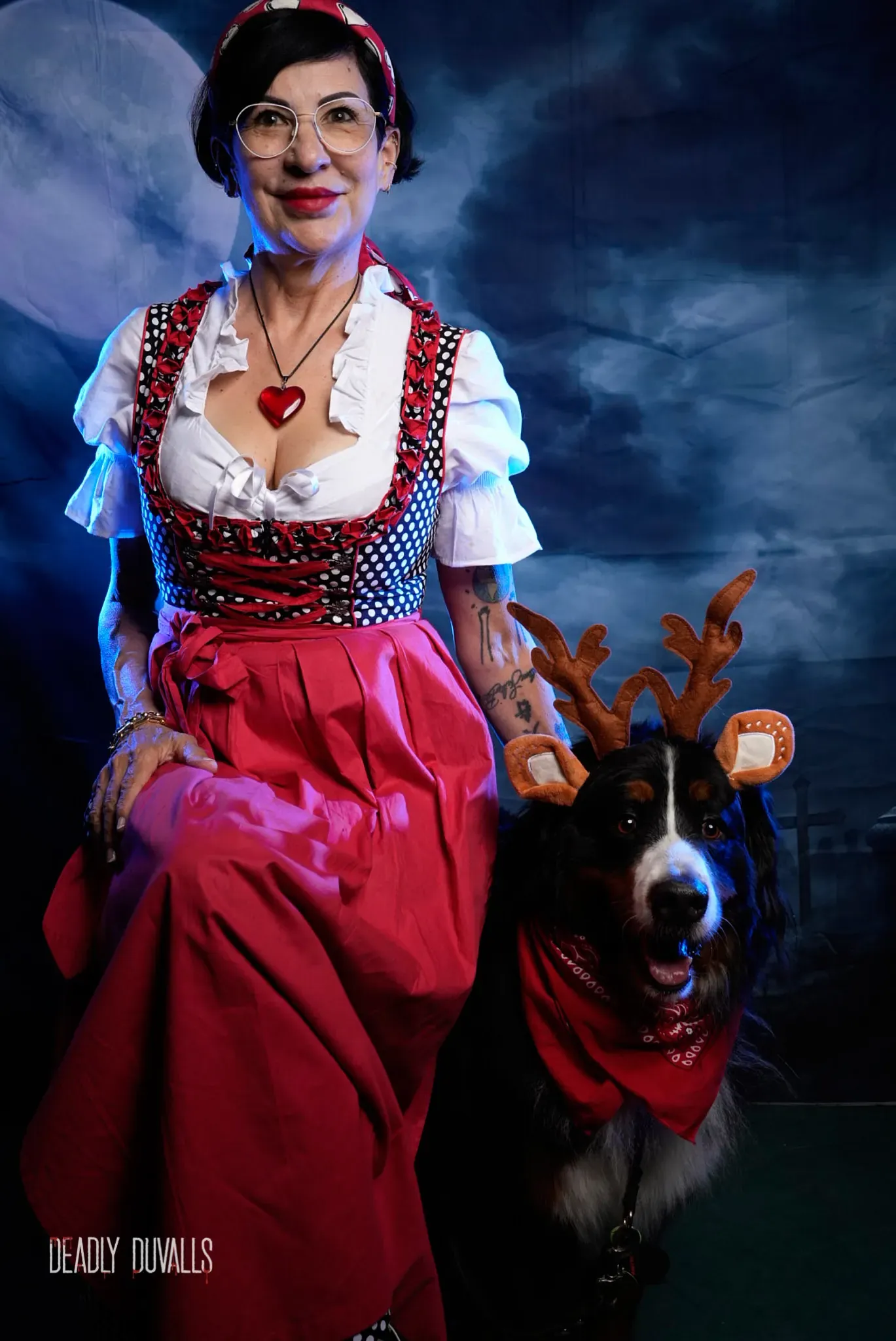 Guest in a red and black dirndl posing with her Bernese mountain dog, who is wearing reindeer antlers and a red bandana, at the Howl-O-Ween photo booth in Los Angeles