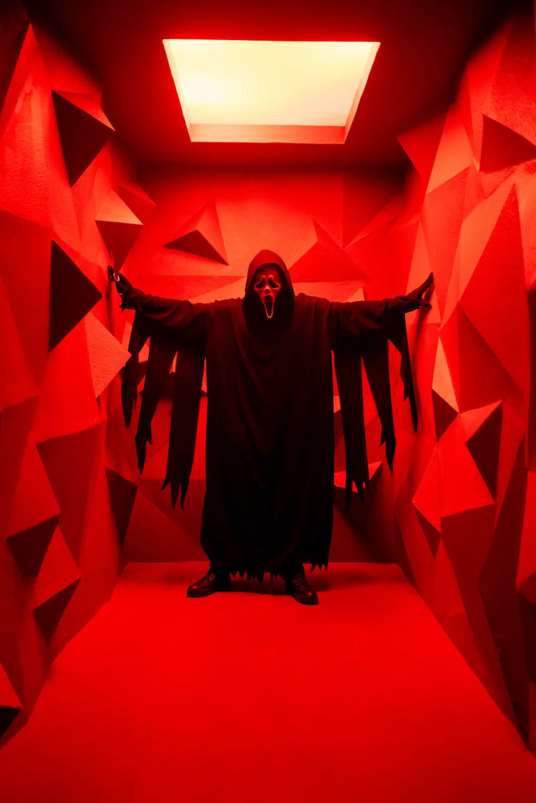 Ghostface with arms outstretched wide in a red-lit corridor of geometric triangles beneath a glowing skylight — narrative horror editorial photography in Los Angeles