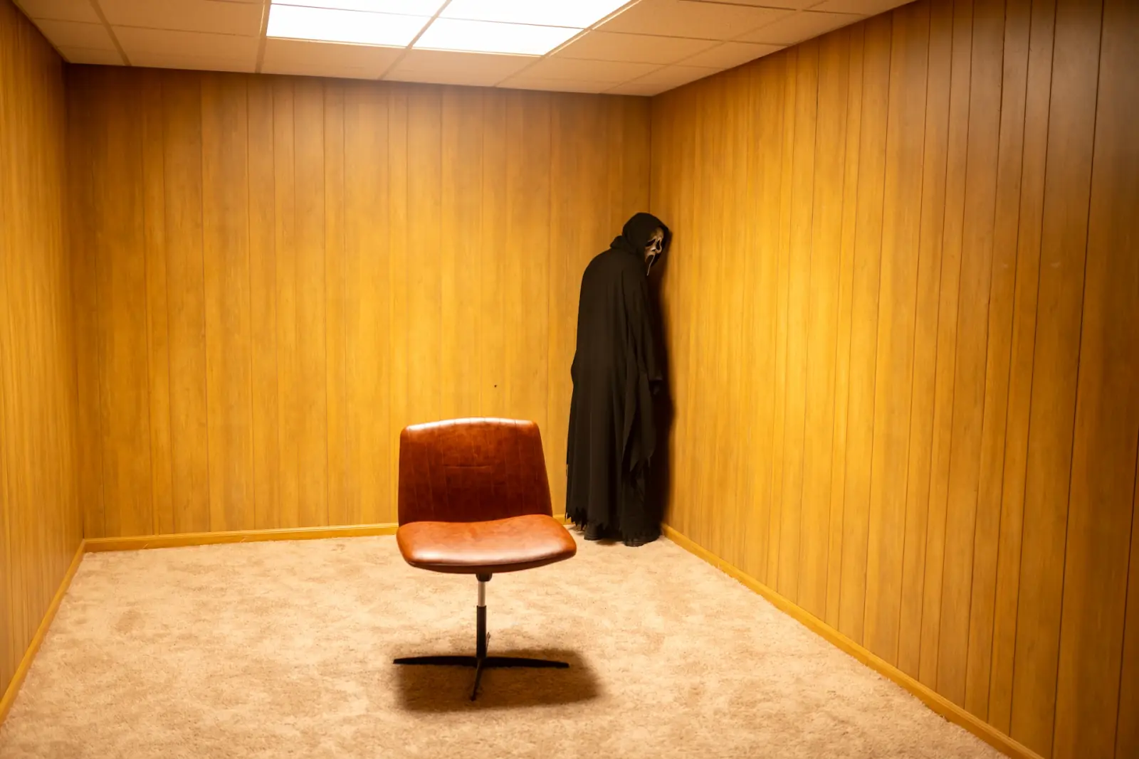 Ghostface facing the corner of a wood-paneled room beside an orange vinyl chair, lit warm and flat — deadpan concept horror photography in Los Angeles