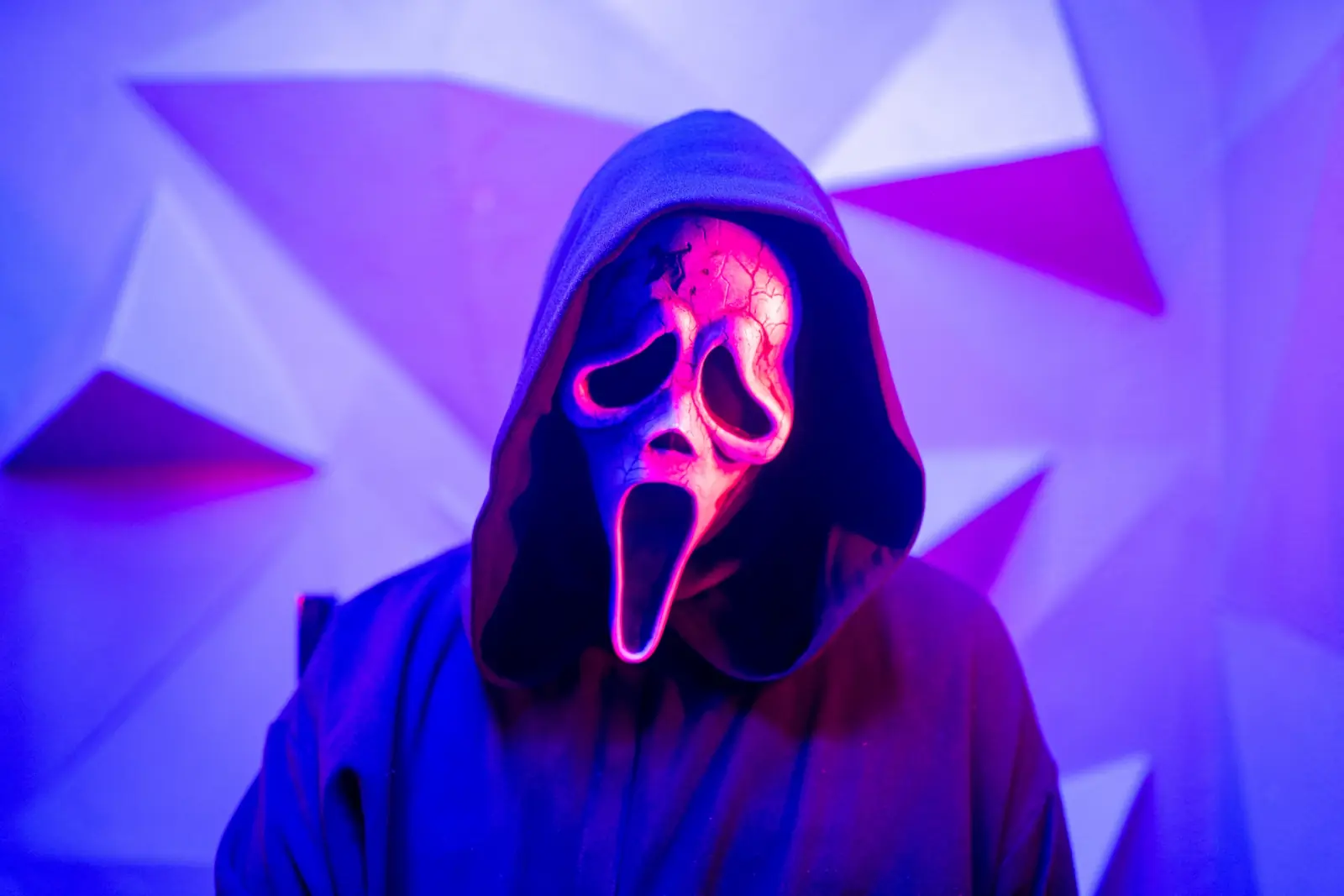 Close-up of the Ghostface mask lit pink against a violet and blue geometric paper backdrop — dual-gel horror portrait photography