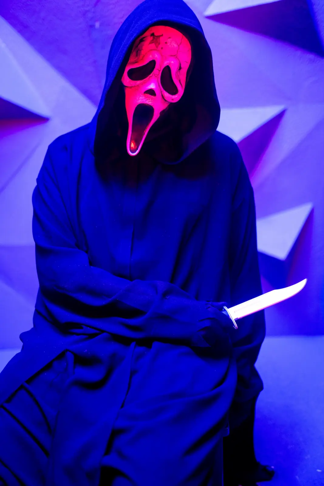 Ghostface holding a bowie knife against a purple and blue geometric backdrop, mask lit hot pink — colour-gelled horror pin-up photography in Los Angeles
