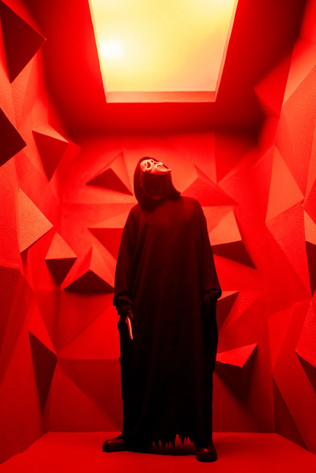 Ghostface tilting their head up toward a glowing ceiling panel in a red room built from geometric triangles, knife at their side — dark editorial horror photography
