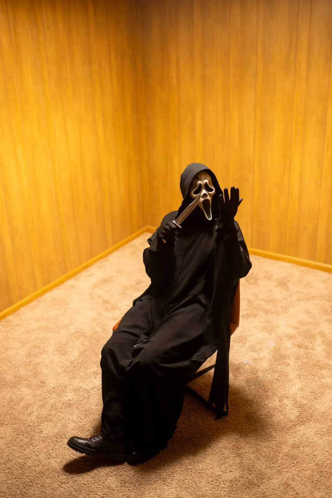 Ghostface seated on an orange chair in a wood-paneled room, raising a knife and waving at the camera — comedic horror portrait by Deadly Duvalls